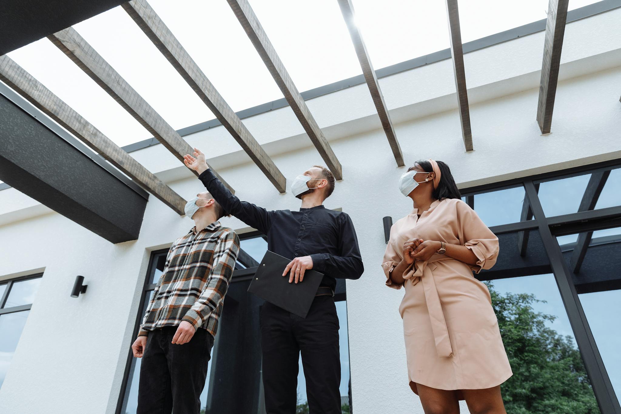 Masked real estate agent showcases modern architecture to clients outdoors.